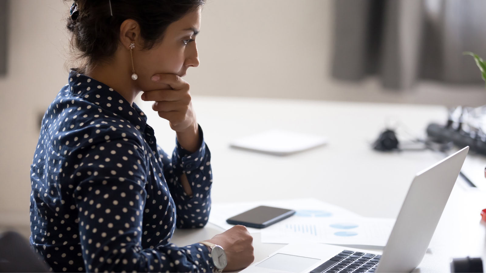 Woman in polka dot shirt sits at a desk, looking intently at a laptop screen with her hand on her chin; MOD Ventures LLC papers and a phone—perhaps reviewing Accounting or Fractional CFO reports—are on the desk.