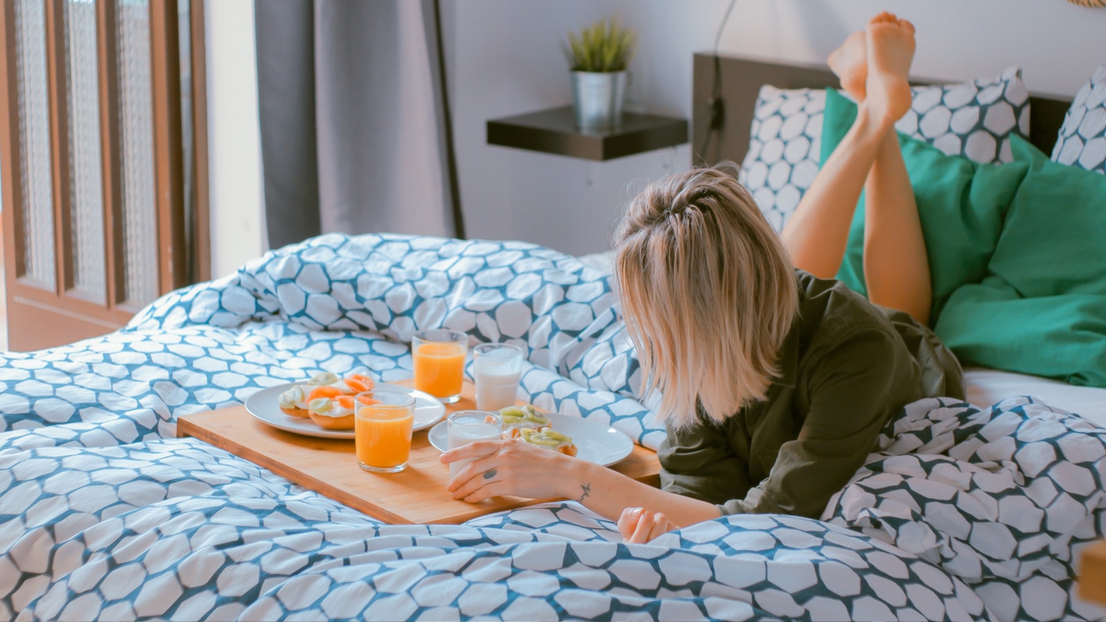 A person lies on a bed with patterned bedding, facing a tray holding plates of food and glasses of orange juice and water, perhaps reviewing Accounting notes from MOD Ventures LLC or tasks from their Fractional CFO.