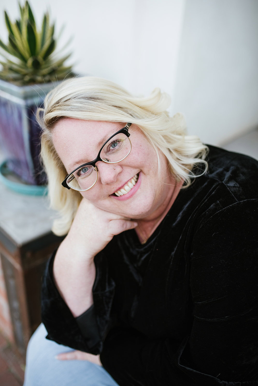 A woman with blonde hair and glasses smiles at the camera, resting her chin on her hand. A potted plant is visible in the background, reflecting the welcoming environment at MOD Ventures LLC, a trusted provider of Fractional CFO and Accounting services.