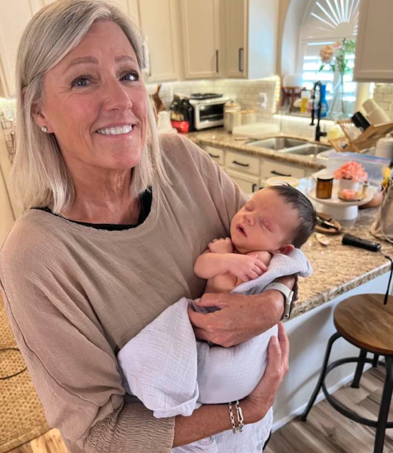 An older woman stands in a kitchen, holding a sleeping newborn wrapped in a white blanket&mdash;a tranquil moment that reflects the care and expertise you&rsquo;d expect from a trusted MOD Ventures LLC Fractional CFO.