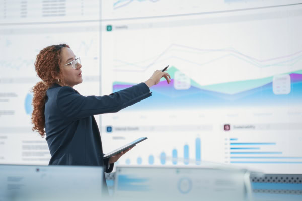 Woman in business attire points at a large digital screen displaying graphs and charts, holding a tablet, illustrating when and how often to use an external financial controller in an office or control room setting.