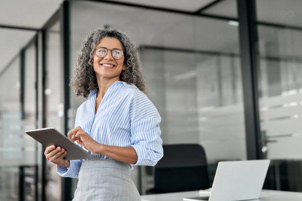 A woman with curly gray hair and glasses stands in an office, smiling and holding a tablet—perhaps reviewing the responsibilities of a fractional controller. A laptop is on the desk beside her.
