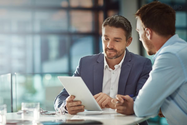 Two men in business attire sit at a table in an office, looking at a tablet and discussing documents related to fractional CFO services.