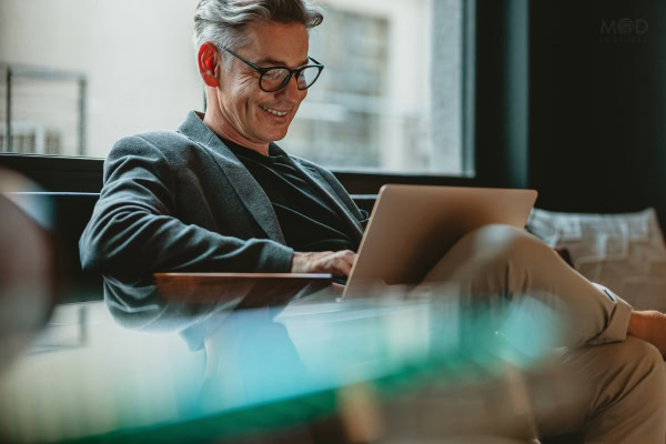 Man with glasses and gray hair sits on a couch, smiling while working on a laptop in a modern indoor setting—perhaps researching how to become a fractional CFO.