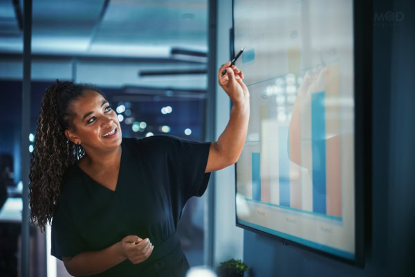 A woman in a black shirt points at a bar graph on a large screen during a business presentation, illustrating how to hire a fractional CFO in an office setting.