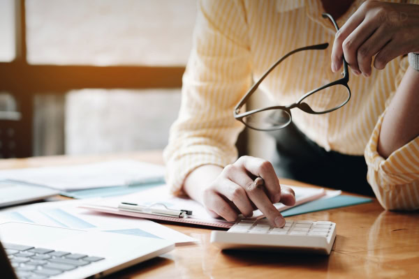 Person in a yellow striped shirt holds glasses and uses a calculator at a desk with documents and a laptop, perhaps reviewing finances—much like what a fractional CFO does. Wondering about the meaning of fractional CFO? They offer expert financial guidance part-time.
