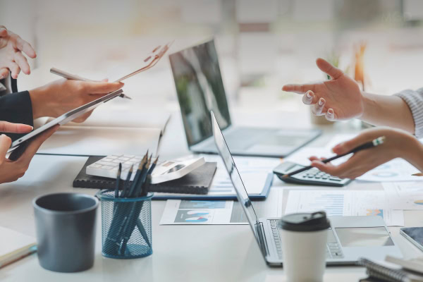People sit at a desk with laptops, documents, charts, and coffee cups, discussing cost structures and streamlining vendor contracts while pointing at papers and screens during a business meeting.