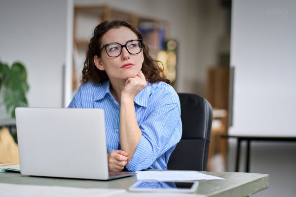 Woman in glasses sits at a desk with a laptop, papers, and tablet, resting her chin on her hand, thoughtfully considering if fractional CFO is worth it for growing her business.
