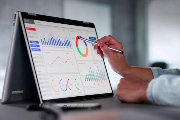 Person points at colorful data charts and graphs displayed on a convertible laptop screen, analyzing business analytics to drive data-backed decisions in a modern, bright office setting.