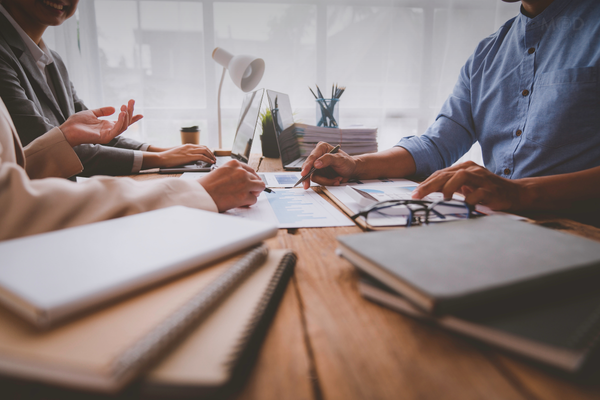 Three people sit at a wooden table in an office, discussing documents and taking notes on cash accounting. Laptops, notebooks, and papers are spread out as they review records to avoid accrual vs cash accounting mistakes.