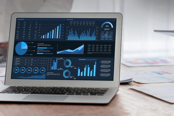 A laptop on a desk displays various data analytics dashboards with charts, graphs, and weekly metrics for the CEO, highlighting numeric statistics and $5M+ performance trends.