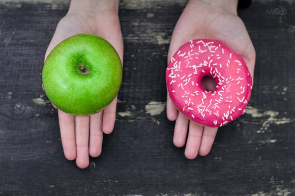 Two hands hold different items, hinting at different outcomes: the left holds a green apple, and the right a pink frosted donut with white sprinkles, both resting on a dark wooden surface—choices any CPA or Fractional CFO might appreciate.