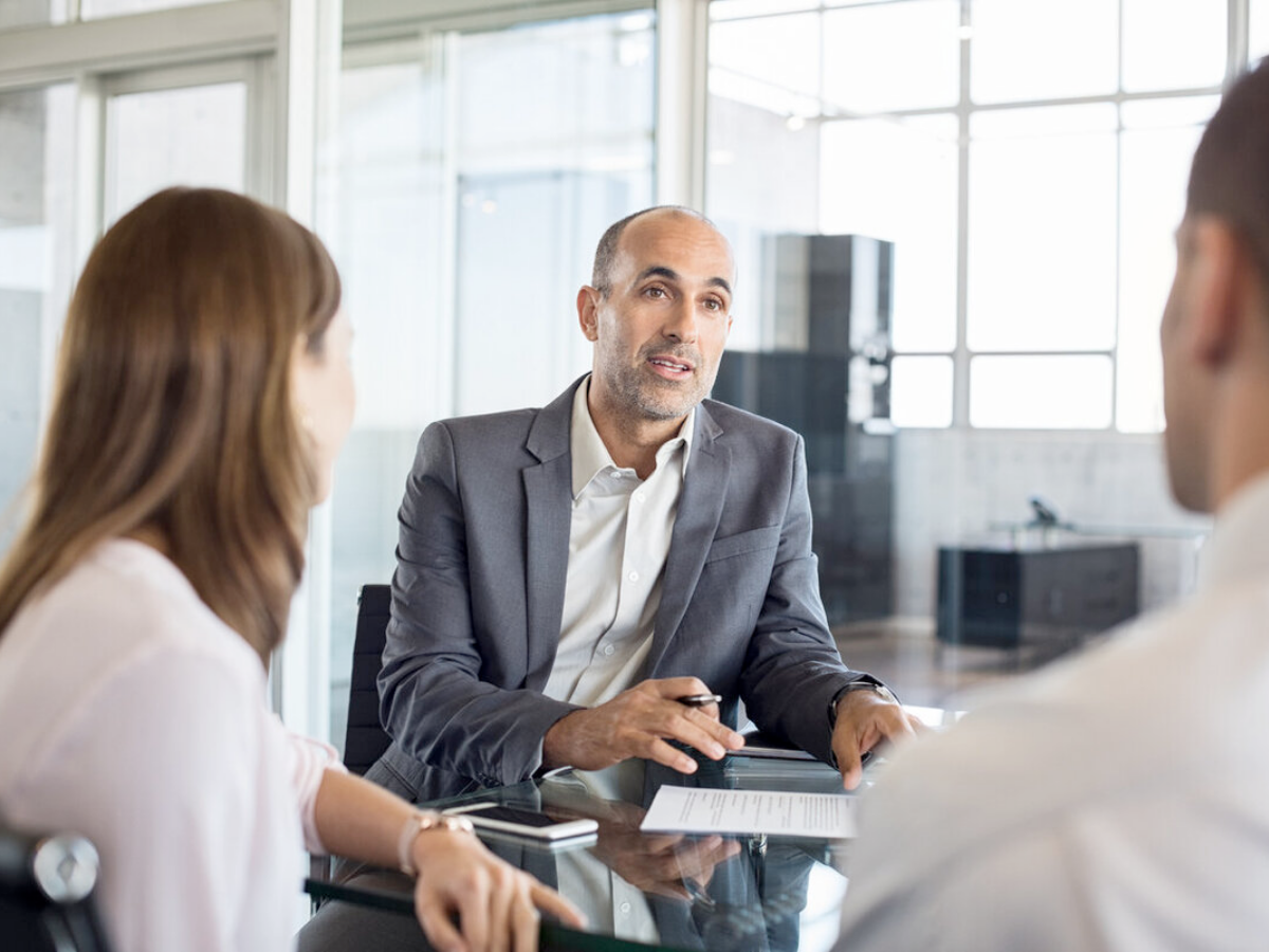 A man in a suit sits at a desk talking to two people, holding a pen and paper, in a modern office—a scene depicting what to expect from a Fractional CFO during the first 30 days.