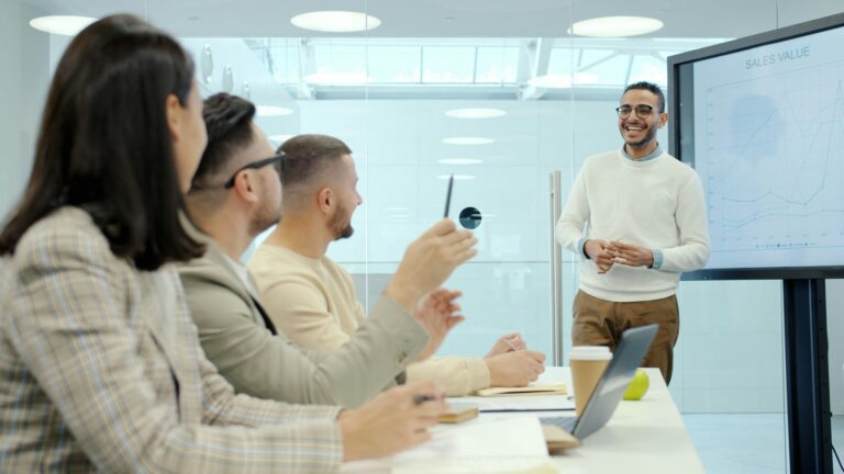 A man presents data on a screen to three colleagues in a meeting room, discussing Controller Services. Notebooks, laptops, and coffee cups are on the table as they explore outsourced controller solutions for their business.