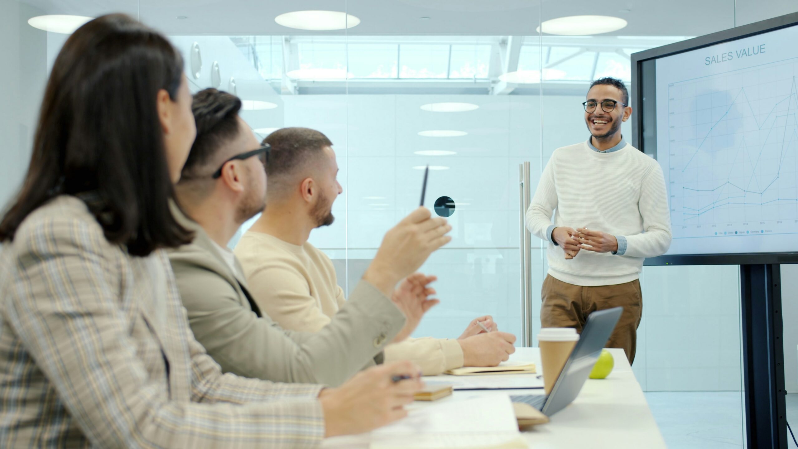 A man presents data on a screen to three colleagues in a meeting room, discussing Controller Services. Notebooks, laptops, and coffee cups are on the table as they explore outsourced controller solutions for their business.