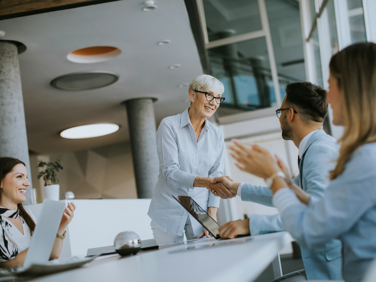 An older woman, the CFO, stands and shakes hands with a seated man at a conference table while two colleagues—likely fellow business owners—smile and clap.