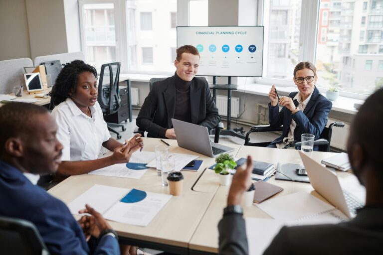 Five people in business attire sit around a conference table discussing, with documents, laptops, and a financial planning cycle presentation displayed on a screen in the background. Their conversation centers on strategies for small businesses to streamline the month-end close process and ensure accuracy by following a comprehensive month-end close checklist.
